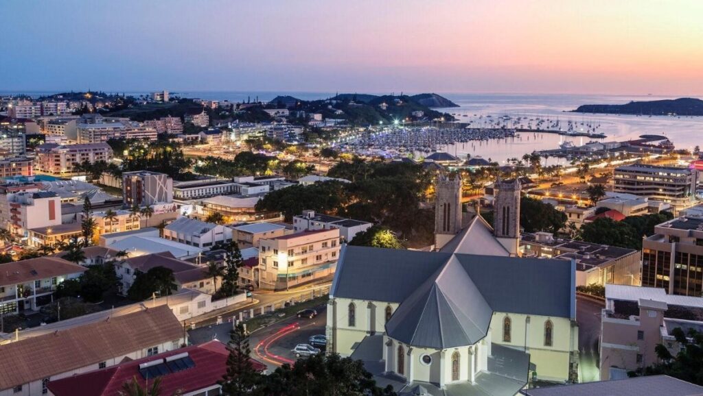 Evening cityscape of Nouméa, New Caledonia’s capital, showing its cathedral, marina, and coastline illuminated under a colorful sunset.