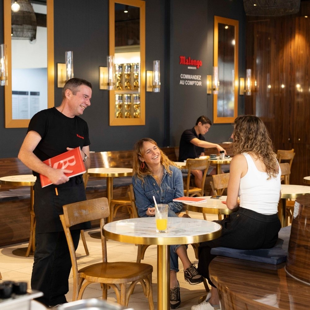 French-style café in New Caledonia with a friendly waiter serving two smiling women at a marble table, blending Parisian charm with island warmth.