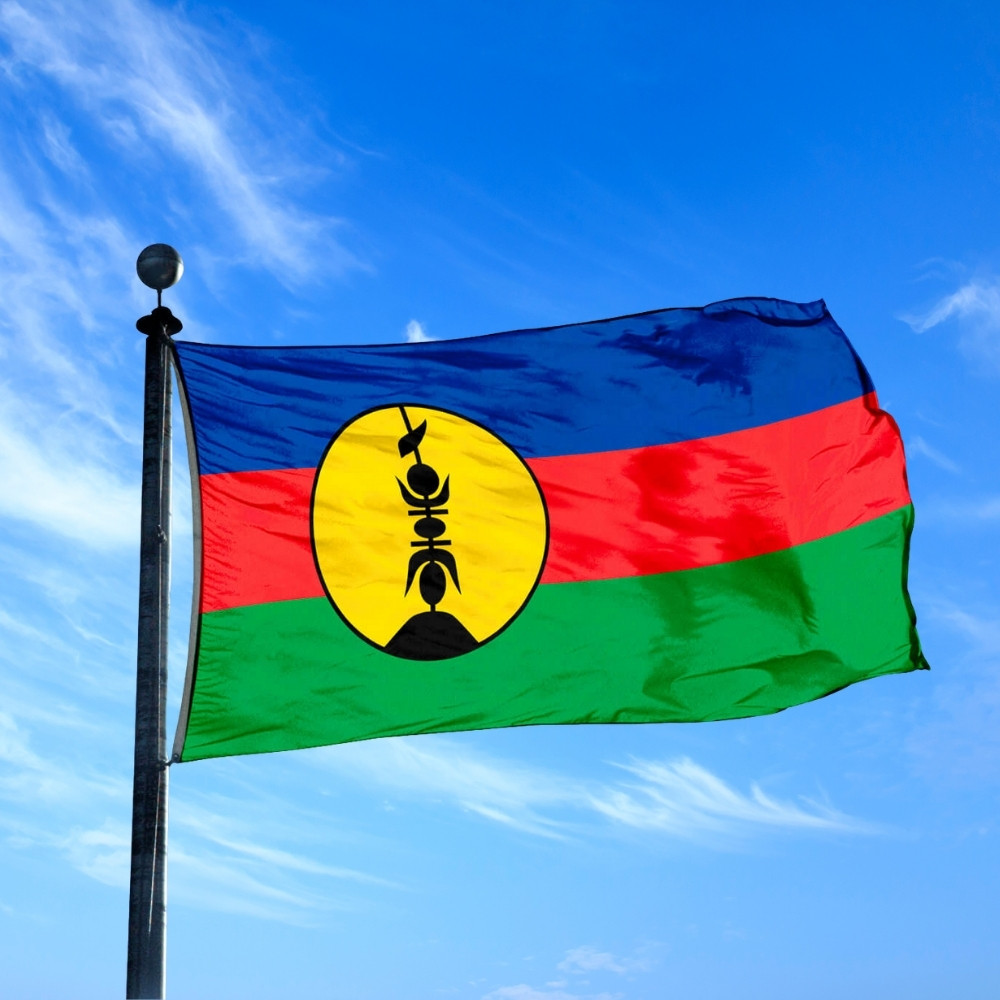 Kanak flag of New Caledonia waving under a bright blue sky, featuring blue, red, and green stripes with a yellow disc and black flèche faîtière symbol.