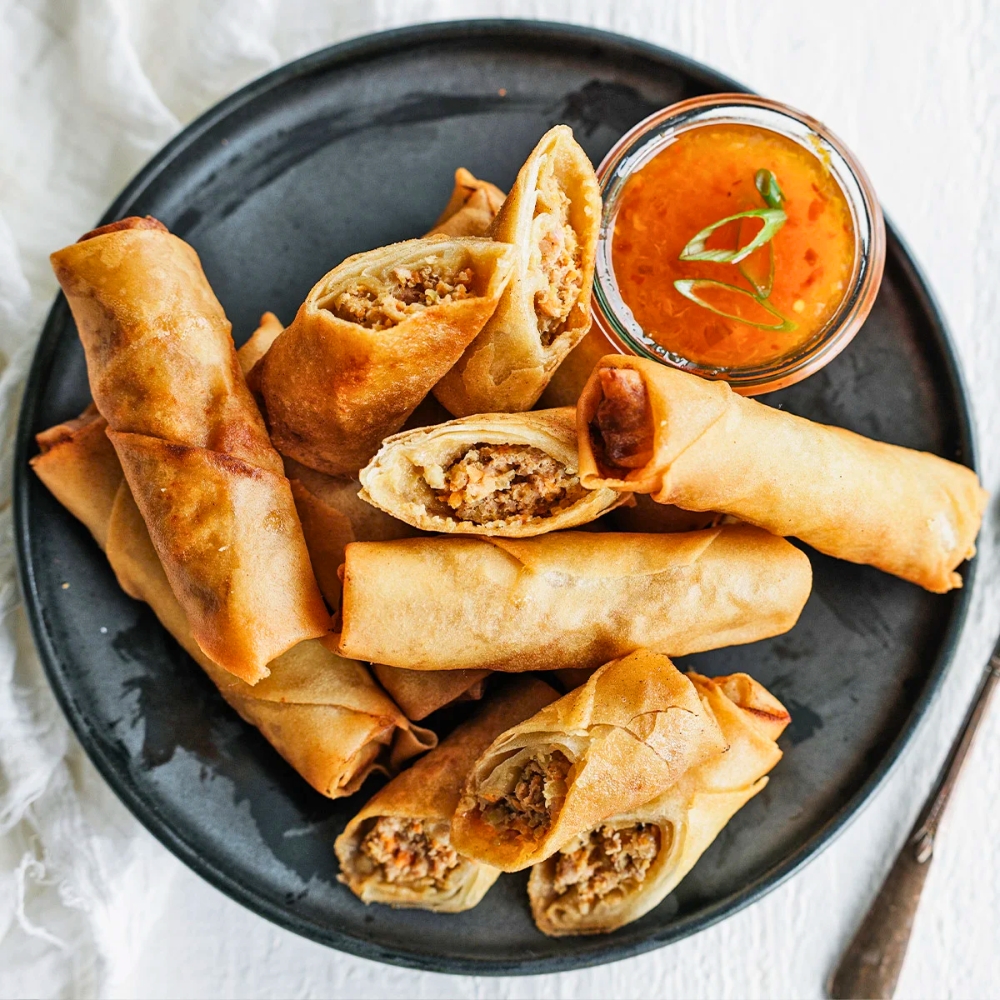 Plate of fried lumpia rolls, some cut open to show the filling, served with a small bowl of orange dipping sauce.