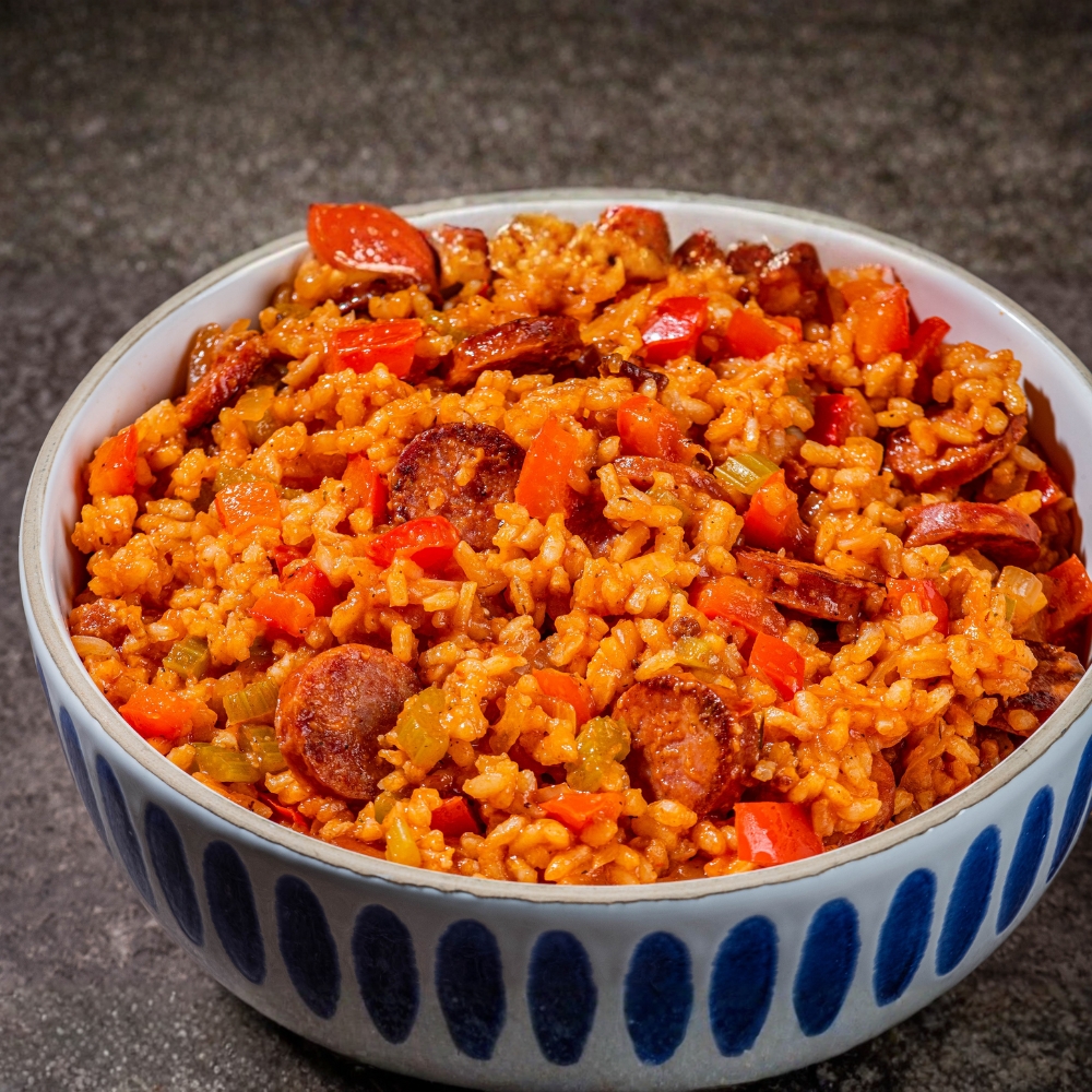Bowl of red rice mixed with sliced sausage and diced vegetables, showing the dish’s bright orange color and texture.