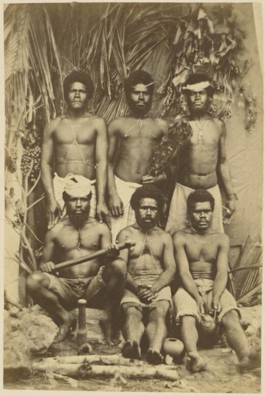 Historic 1870 portrait of six Kanak men in traditional attire surrounded by cultural artefacts, photographed in New Caledonia by W. & E. Dufty.