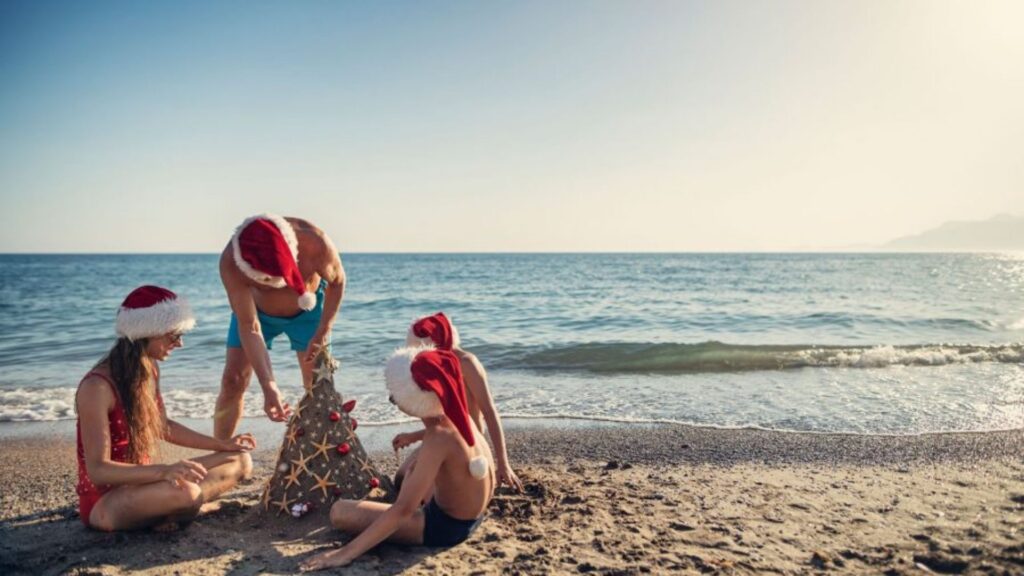 People wearing Santa hats building a small Christmas tree structure on a sunny beach in New Zealand during the summer holiday season.
