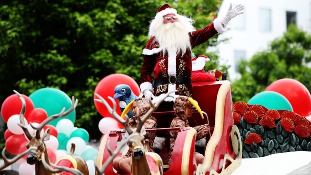Santa riding on a festive parade float in New Zealand, surrounded by colourful balloons, reindeer decorations, and summer greenery.